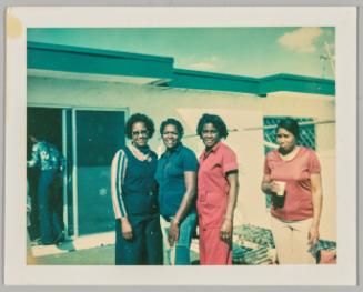 [Three women standing together outside and smiling, another woman standing off to the right and pouting with a cup in her hand, man walking through sliding doors on left ]