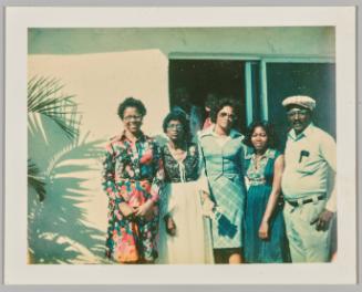 [Four dressed up women and a man in a hat on far right standing together outside in front of sliding glass doors, palm leaves to the left]