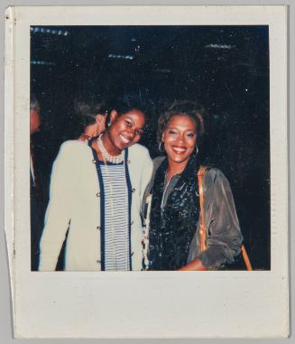 [Two women standing side by side smiling, woman on left wearing baby blue cardigan and striped dress]
