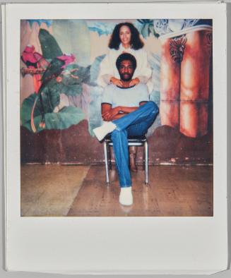 [Woman standing behind seated man with her hands on his shoulders in front of mural on back wall]
