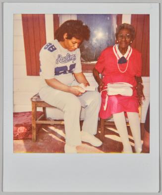 [Elderly woman in red and white outfit sitting on porch with woman sitting next to her holding piece of paper]
