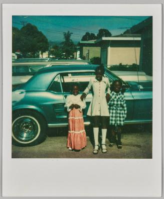 [Three little girls in dresses posing in front of blue car]
