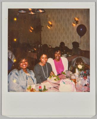 [Three women sitting at a table in front of a birthday cake]
