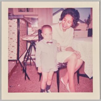 [Little boy wearing a short-suit and bowtie, standing next to woman sitting at table]
