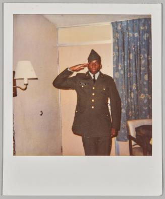 [Young man saluting in military uniform in home] 
