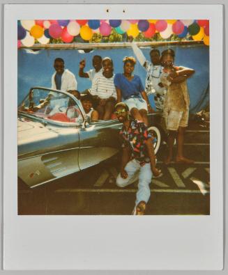 [Group posing in and around car, colourful balloons floating above them]
