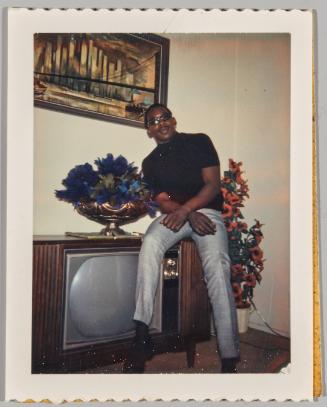 Good things come to those who wait [Man leaning on TV stand wearing grey pants and sunglasses]
