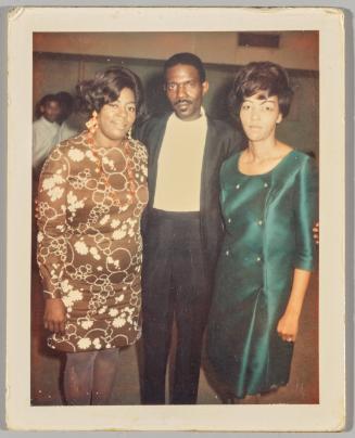 [Man posing with arms around two women in formal apparel with man in white in background]
