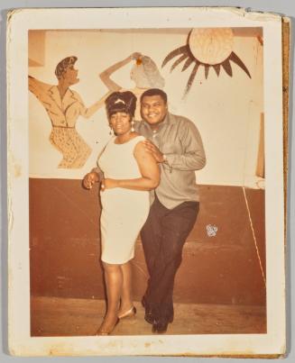 [Man and woman posing in front of mural of people dancing] 
