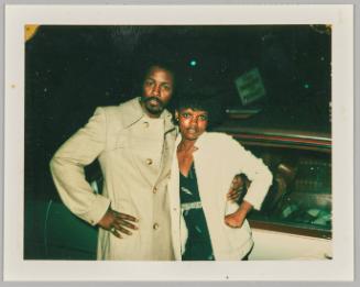 [Man and woman posing with arms around each other and hands on hip in front of car]
