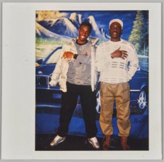 [Two men posing in front of mural of car with mountains]
