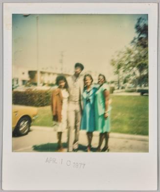 [Three women and a man posing in parking lot in front of grass]