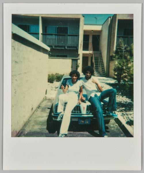 [Young woman and man wearing bellbottoms sitting on roof of car, woman giving thumbs up]
