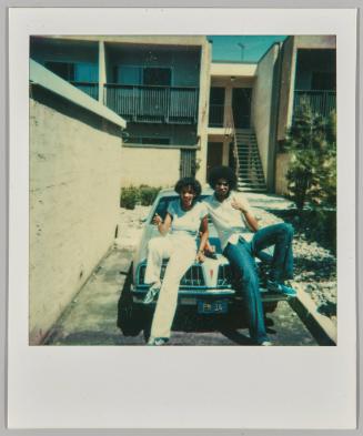 [Young woman and man wearing bellbottoms sitting on roof of car, woman giving thumbs up]
