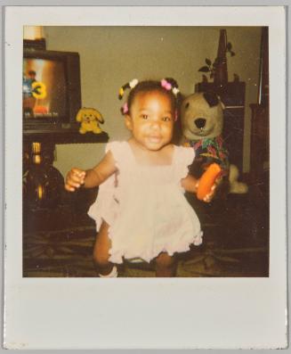 8-91 11 1/2 Month Los Andrews [Toddler standing with braided hair holding a carrot]
