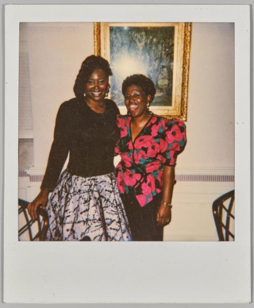 [Two women dressed formally posing in front of embellished framed painting]
