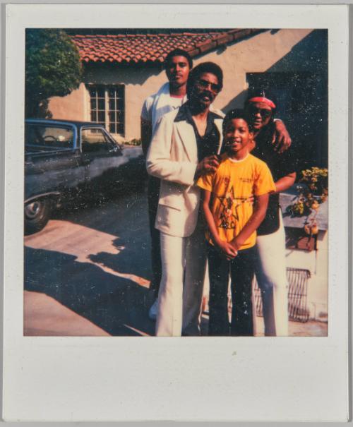 [Man, woman, adolescent boy and young boy posing in front of house and car]
