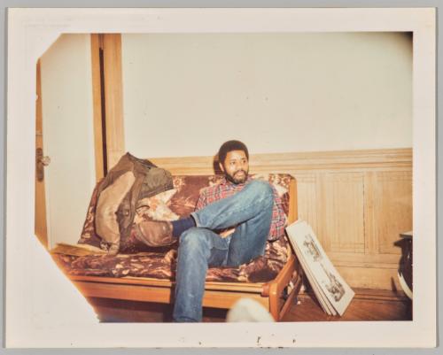 [Man seated crosslegged on couch with art prints leaning against couch]
