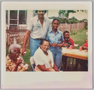 [Two elderly women in folding chairs in front of standing man and young woman in backyard with pop cans on table]
