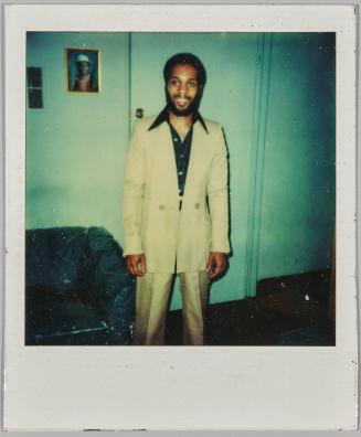 [Man in pale yellow suit smiling in front of door]
