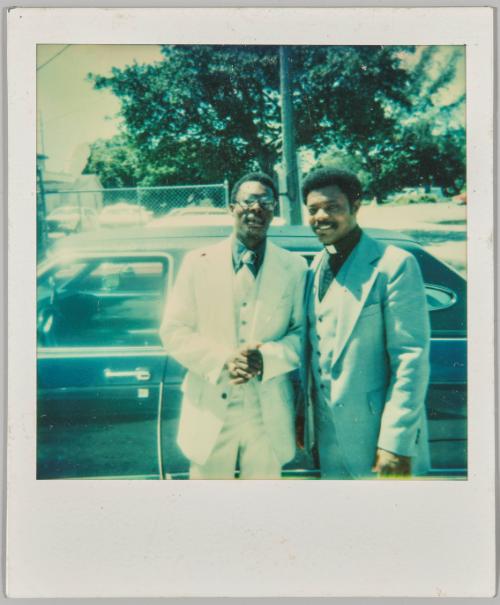 [Two men dressed formally standing beside car, man on left is a priest]
