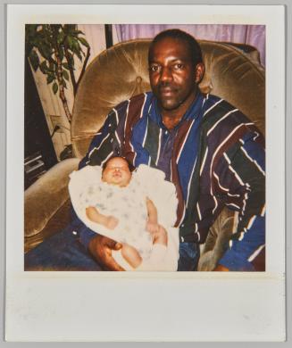 [Man in striped dress shirt sitting in chair holding a sleeping infant]