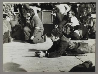 [A man laying down, holding a firearm, many around him seeking shelter from the nearby danger]
