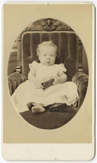 [Seated baby in armchair, holding one shoe and wearing another]