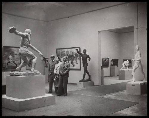 Three men (with Nazi insignias) inside a gallery - exhibiting sports themed paintings and sculptures.