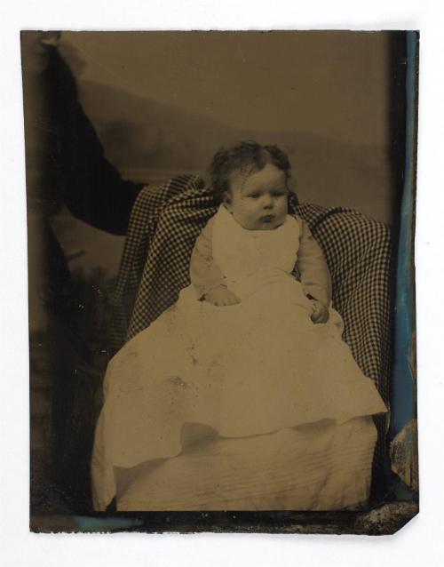 Portrait of baby girl in long white dress (seated on covered chair, supported by cropped figure)