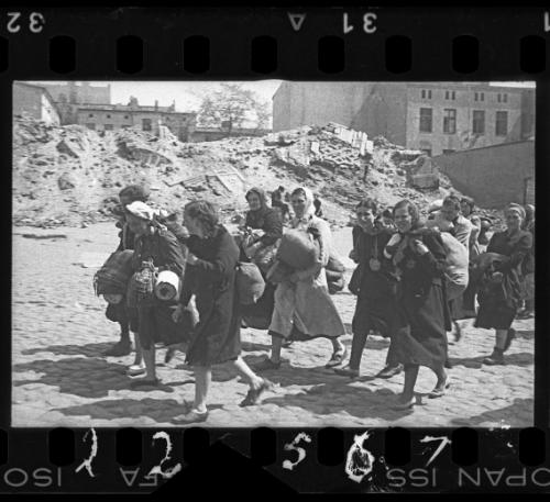 Group of women with sacks and pails walking past synagogue in ruins heading for deportation